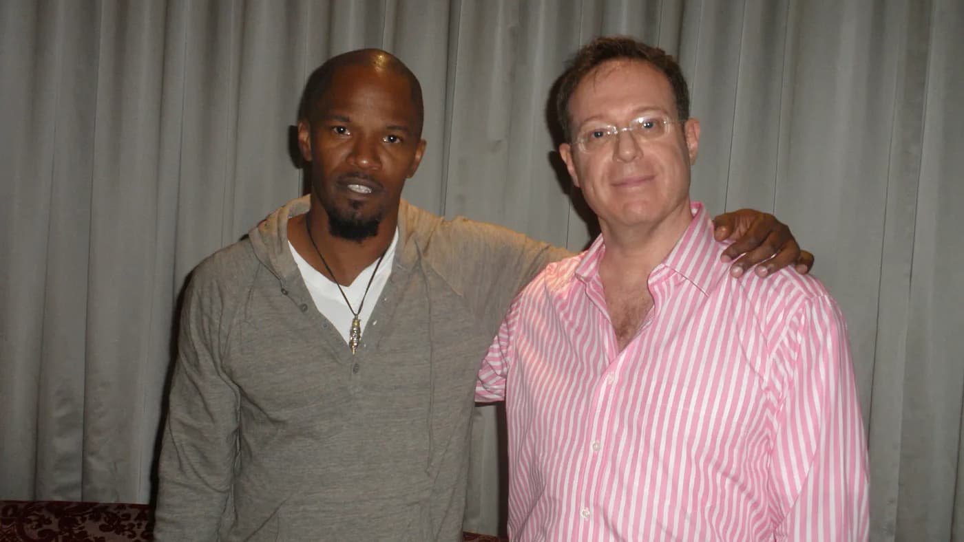 a photo of ami shafrir and jamie foxx standing in front of a grey curtain, they have a hand on each others shoulder