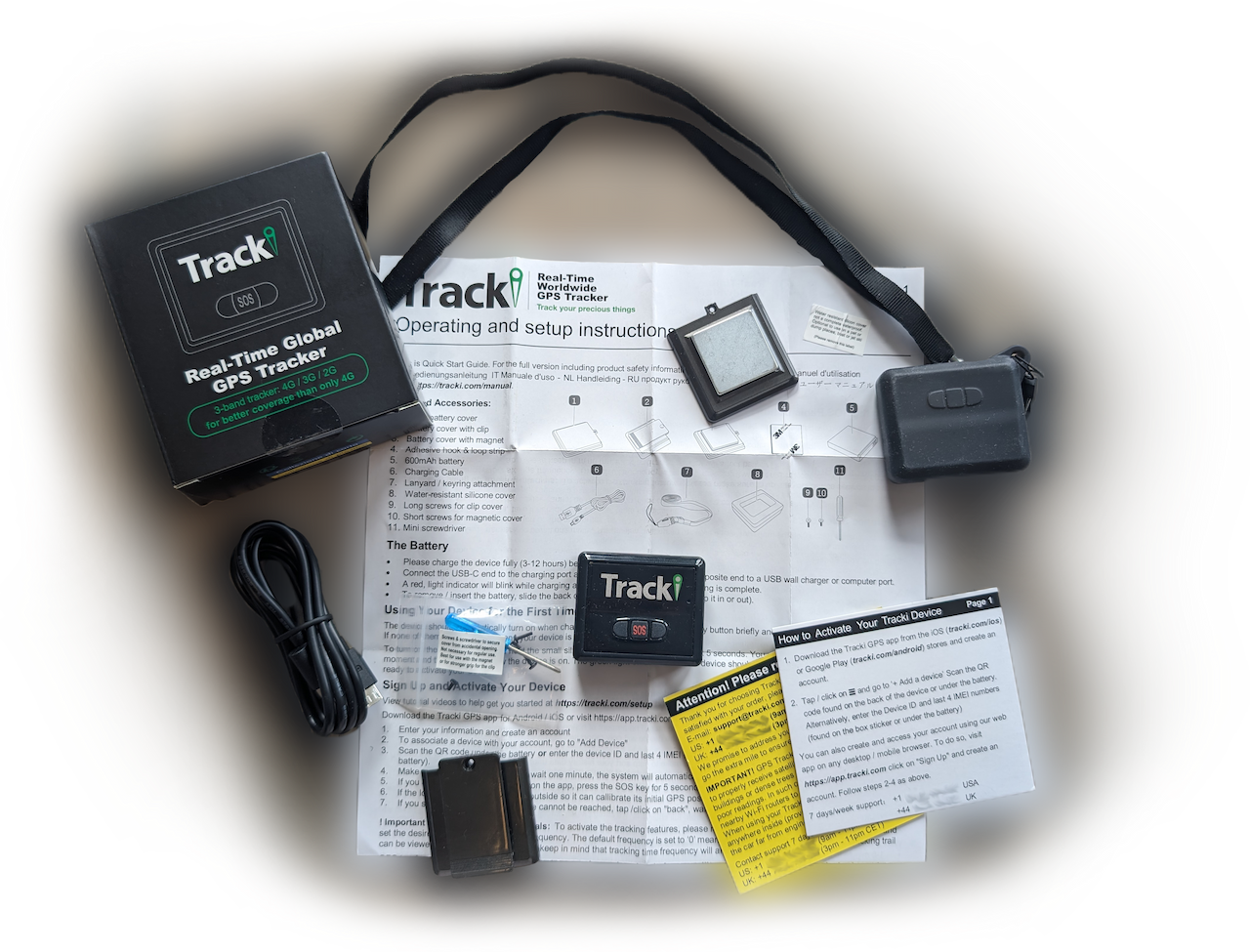 a photo of the tracki device on a table with all other shipment contents spread out around it. those include the black and green box itself, a charging cable, a tiny screwdriver and some screws, setup instructions, two small cards with additional instructions, support phone numbers, and multiple attachments and covers for the device.